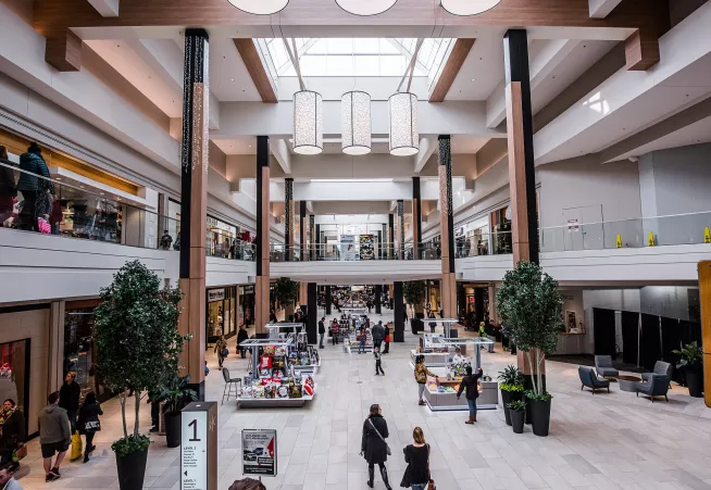 open mall with two floors, skylights creating a sunlit atmosphere, people walking in and out of stores